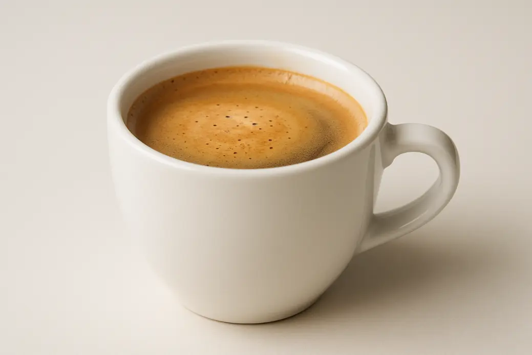 A white coffee cup containing coffee with a visible crema layer, photographed from a 45-degree angle and centered on a neutral background